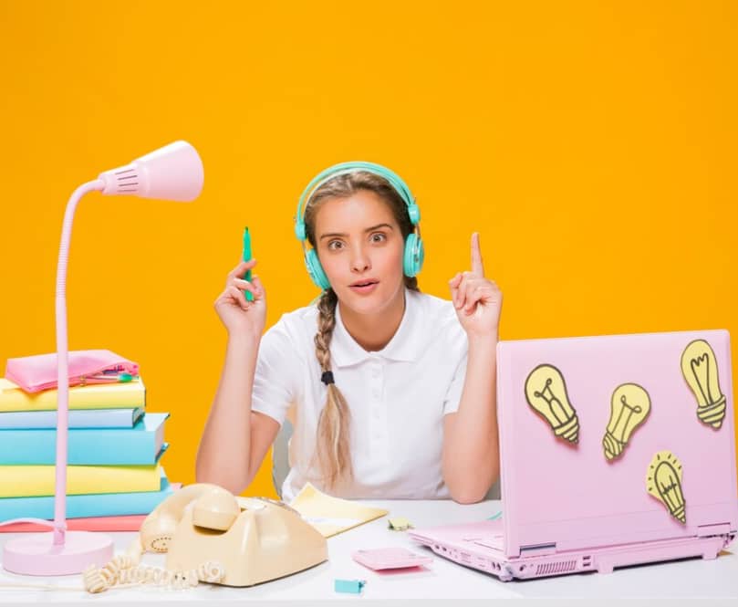 A surprised girl with headphones sits by a pink lamp and laptop